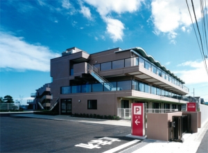 介護施設の建物外観。3階建てのモダンな設計で、駐車スペースと入口が見える。