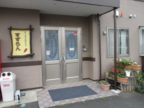 介護施設の玄関入口。二重引き戸で、左側に黄色い看板、右側に植栽がある。